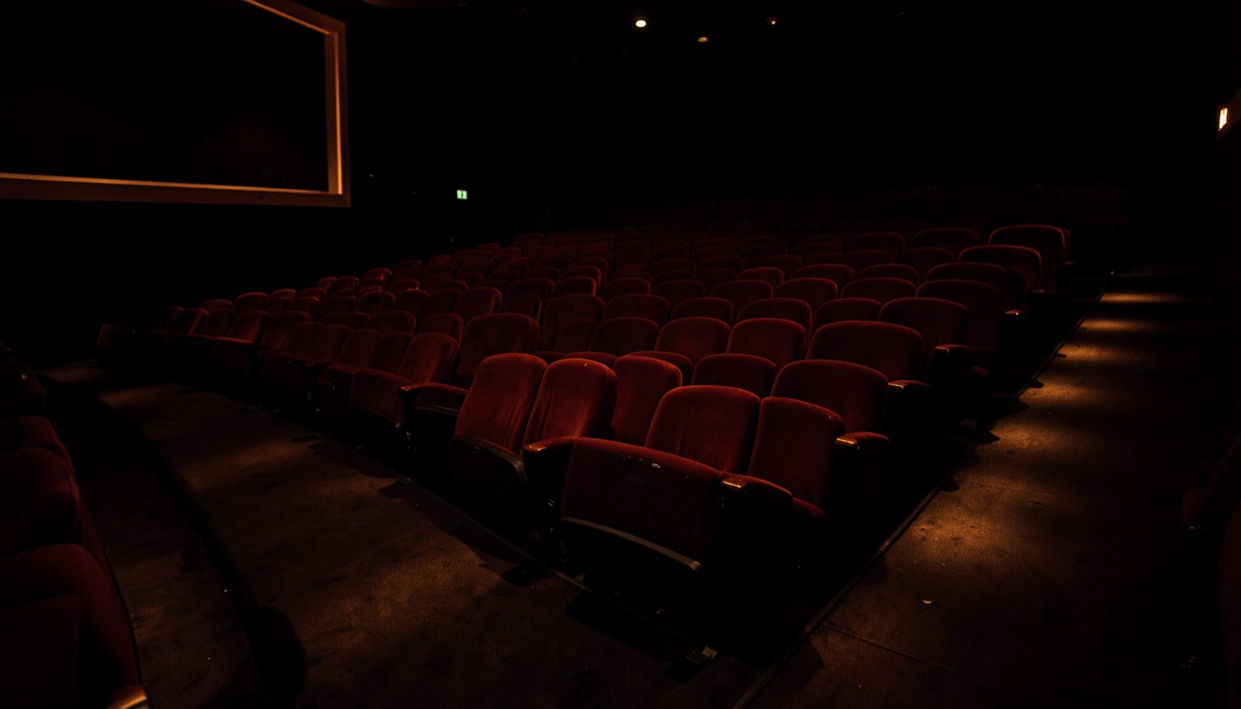 Dark cinema auditorium with rows of empty red velvet seats, soft amber aisle lighting casting warm pools on the floor, deep shadows throughout