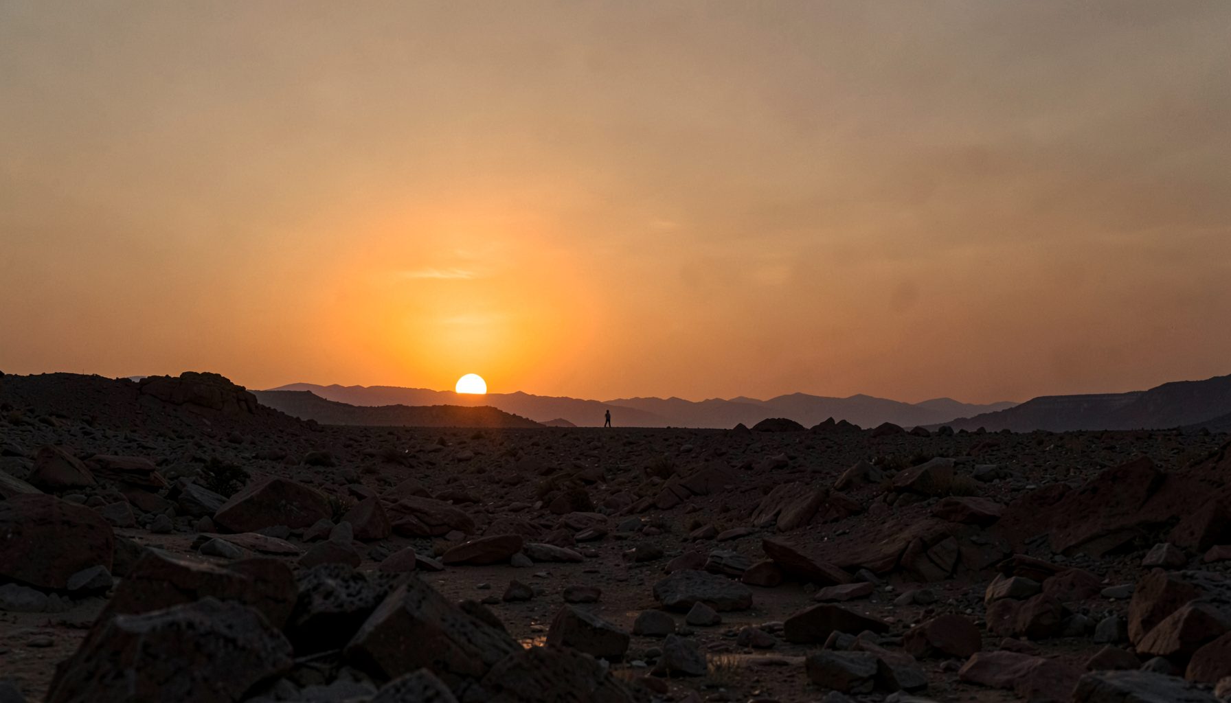 Distant twin suns setting over a rocky desert landscape, warm amber and dusty orange sky, lone figure silhouette on the horizon