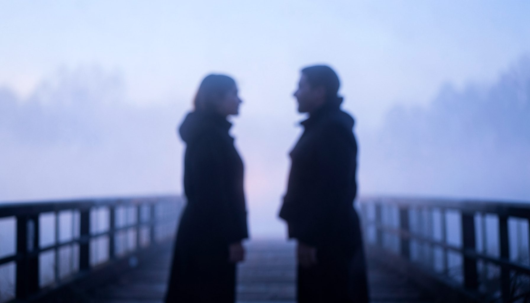 Soft-focus scene of two silhouetted figures facing each other on a misty bridge at dawn, pale blue and lavender tones
